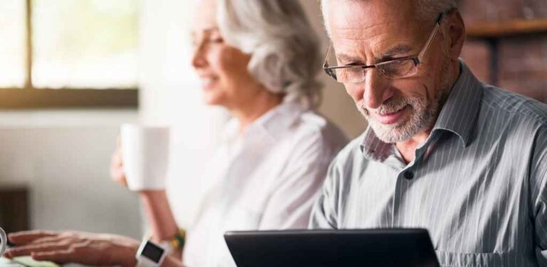 Older man looking at laptop with older woman in background